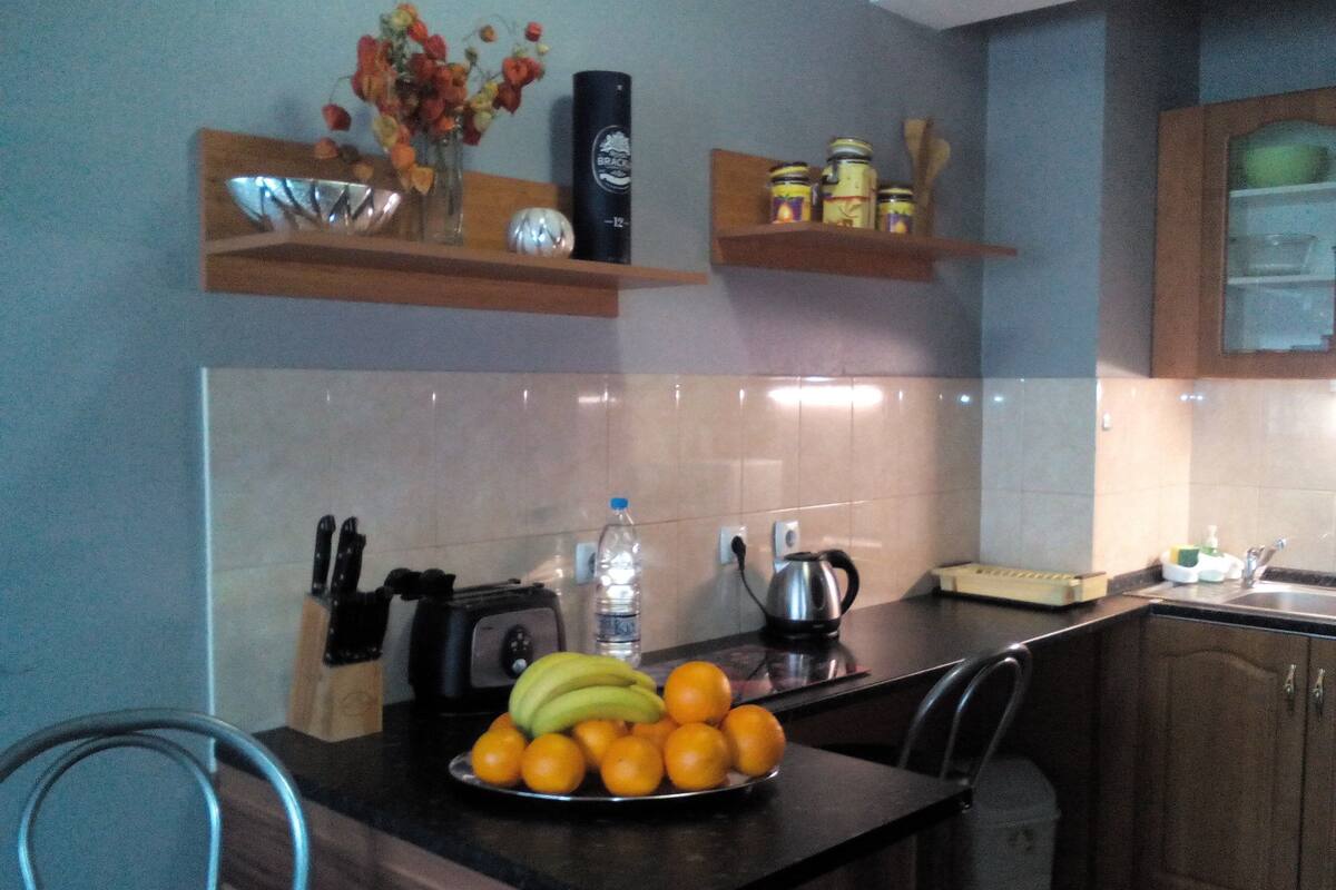 A kitchen with a wooden table, a fruit bowl, a kettle, and a water bottle.