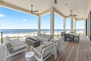 A covered outdoor deck with a view of the beach, featuring a sofa, chairs, a table, and a fire pit.