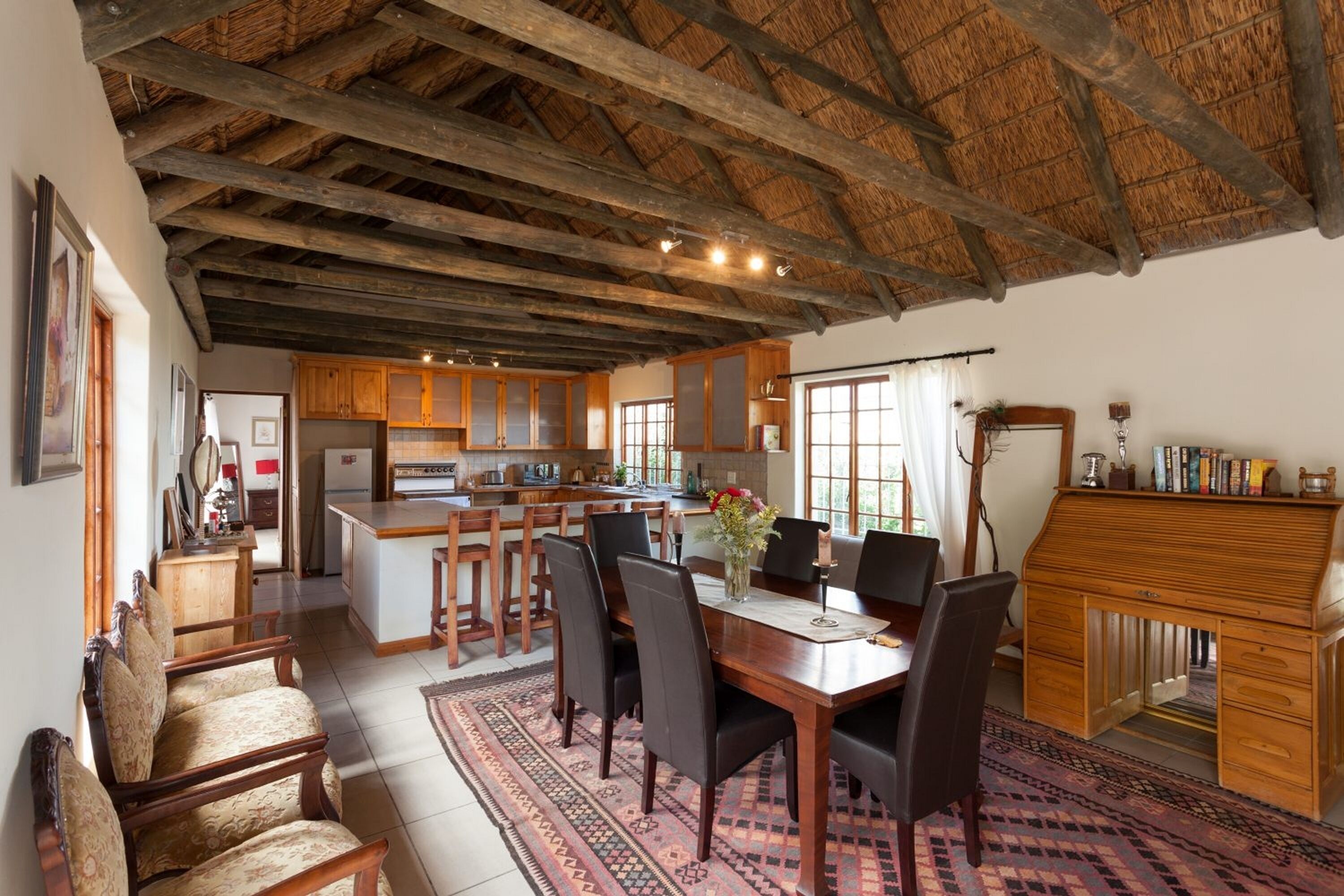 A spacious dining area with a wooden table and chairs, a kitchen in the background, and a rustic ceiling.