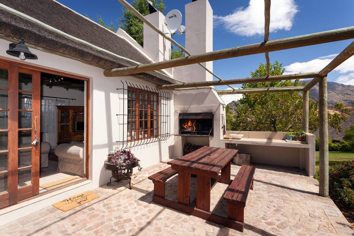 Un patio con una mesa y bancos de madera, una parrilla y una puerta que da a una habitación con cama y cortinas.