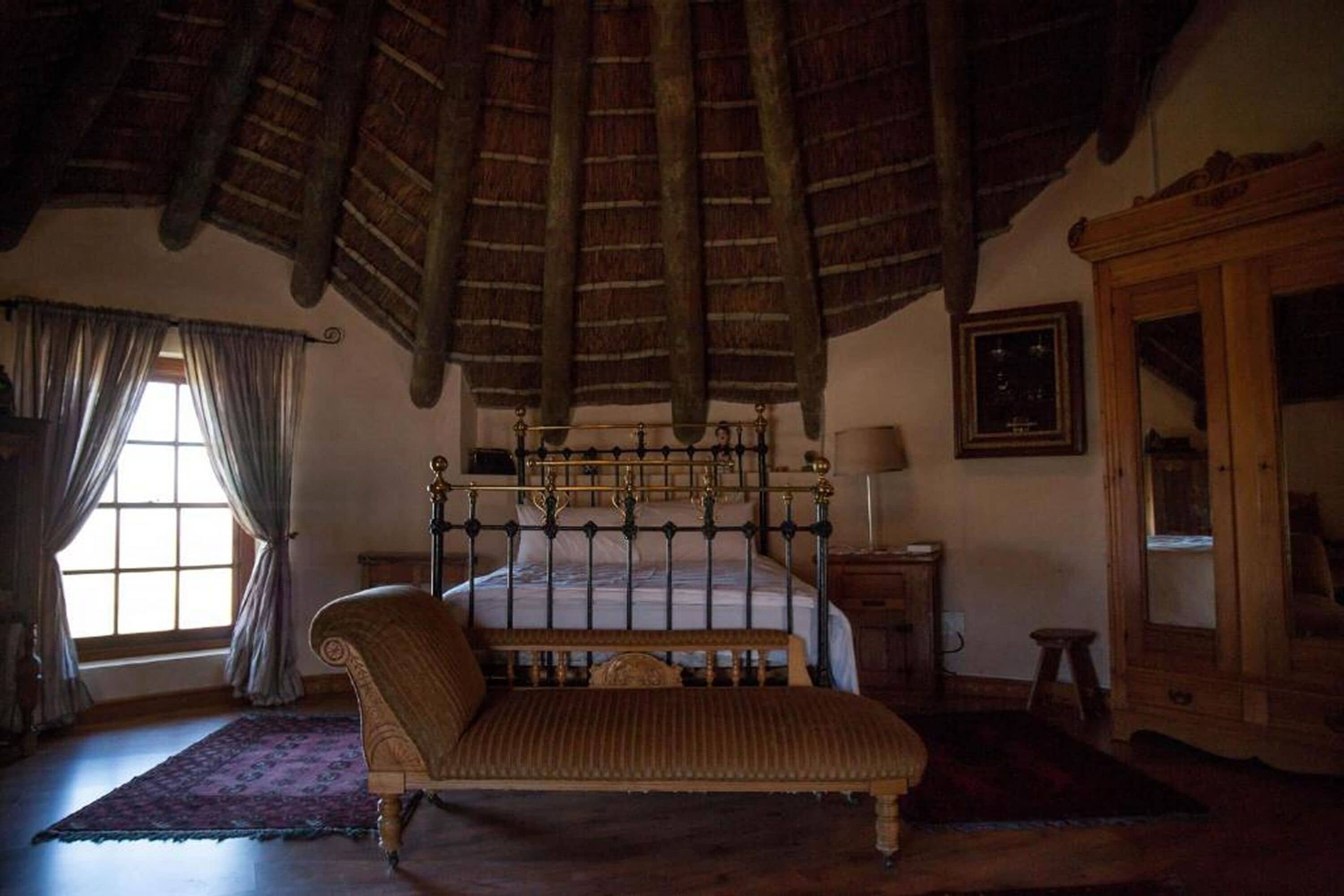 A bedroom with a four-poster bed, a wooden bench, a wooden cabinet, and a large window with curtains.
