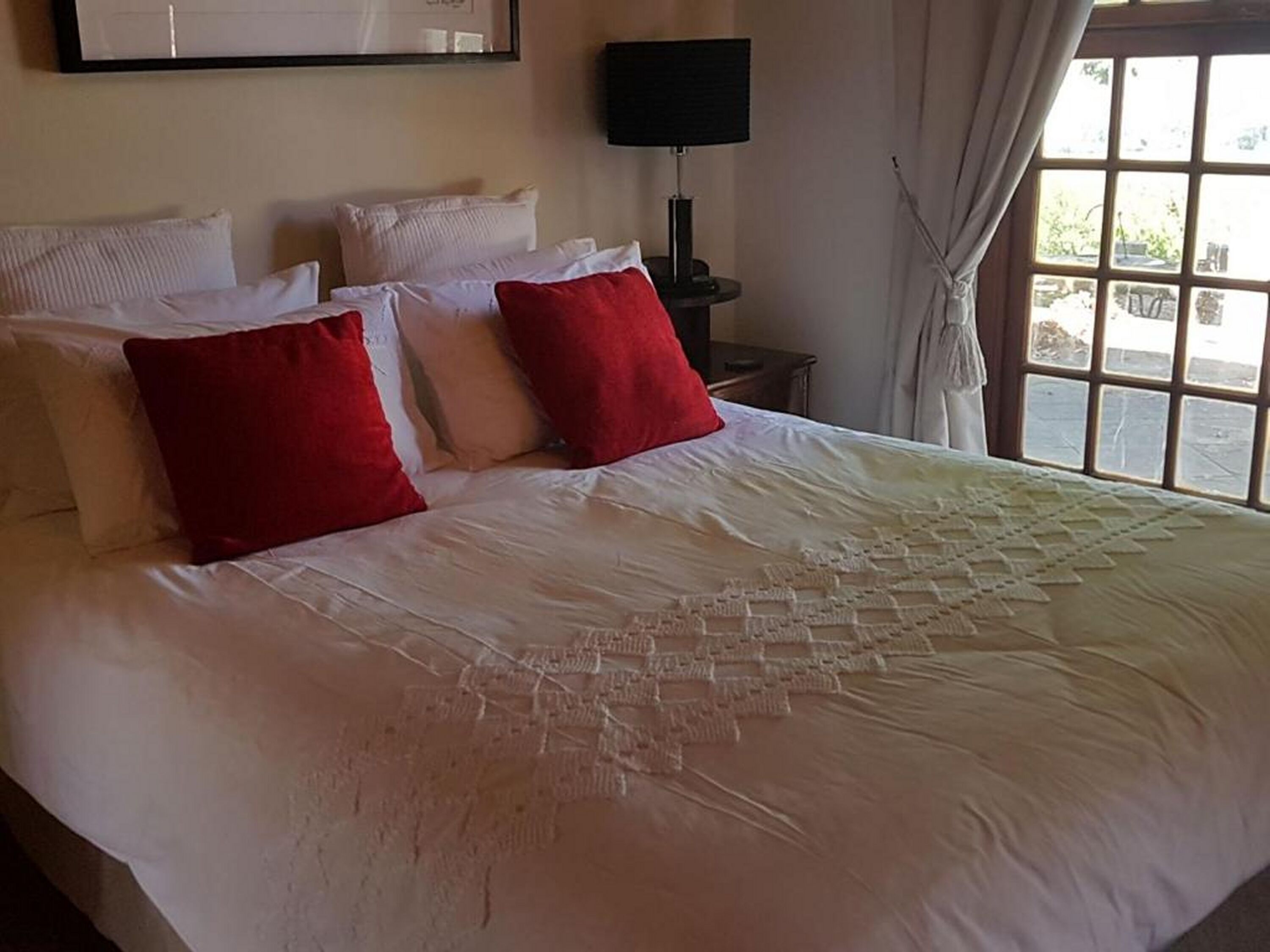 Una cama con almohadas blancas y rojas, una mesita de noche con lámpara y una ventana con cortinas.