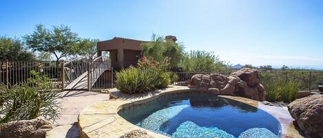 A swimming pool with a stone border, surrounded by rocks and plants, and a small building with a balcony.