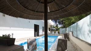A pool area with a thatched umbrella, lounge chairs, and a white table. - Hotel San Domenico (Cervia)