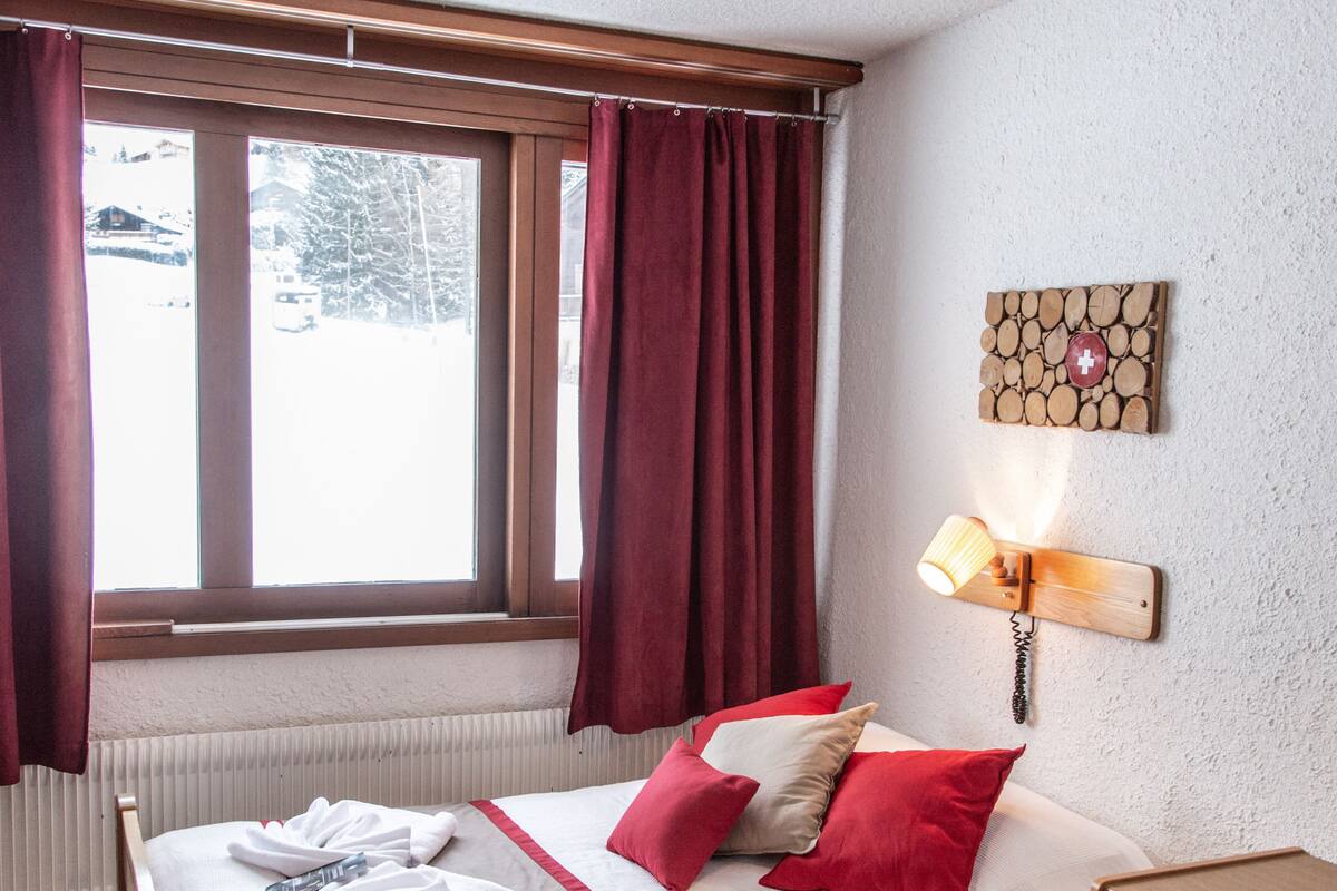 Una habitación de hotel con cama, mesita de noche, lámpara, ventana con cortinas y un aplique en la pared.