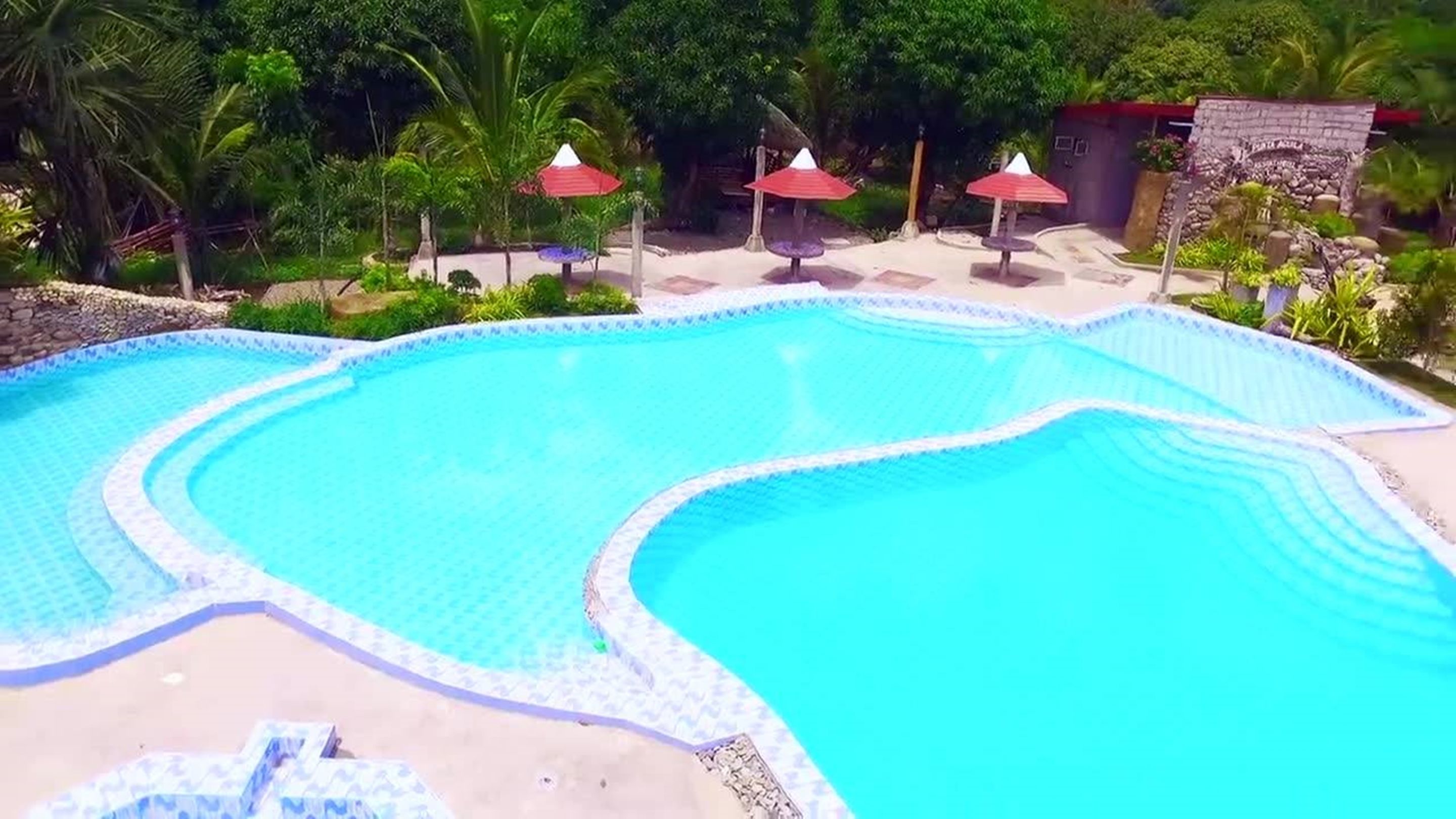 A swimming pool with a wavy edge design, surrounded by a sandy area and red umbrellas.