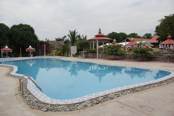 Una piscina con borde de azulejos, rodeada por un marco de piedra. Se observan varias construcciones pequeñas con tejados rojos, posiblemente glorietas, y una variedad de plantas y árboles al fondo.