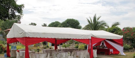 A tented outdoor event space with tables and chairs, surrounded by greenery.