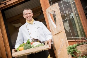 A chef holding a wooden tray with fresh vegetables, standing in front of a wooden door with a heart-shaped cutout. - Gasthaus-Pension Zum Kreuz (Stuehlingen)