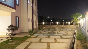Vista noturna da fachada de um edifício, com um pátio pavimentado e uma pequena árvore.