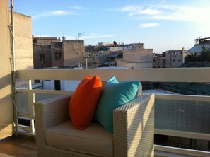 A wicker sofa with cushions on a balcony overlooking a cityscape.