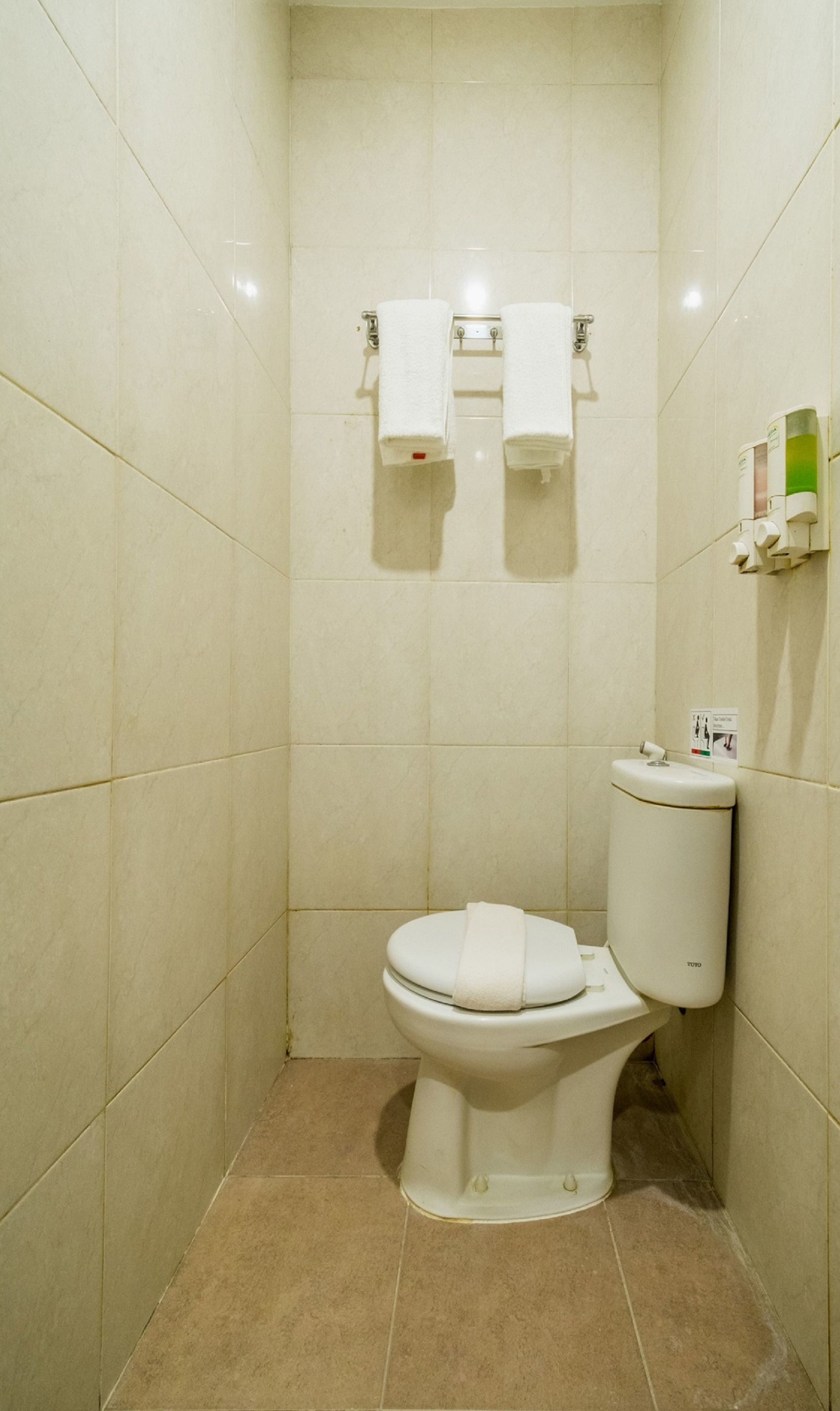 Une petite salle de bain propre, avec des toilettes, deux serviettes blanches suspendues à un porte-serviettes et un support à papier toilette fixé au mur.