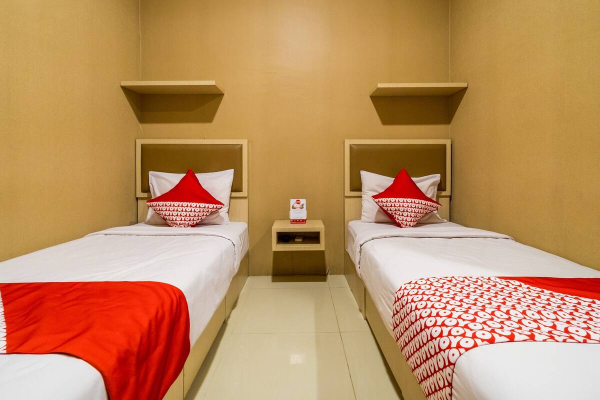 A hotel room with two beds, each with a red and white patterned bedspread, a small bedside table with a tissue box, and a wall-mounted shelf above each bed.