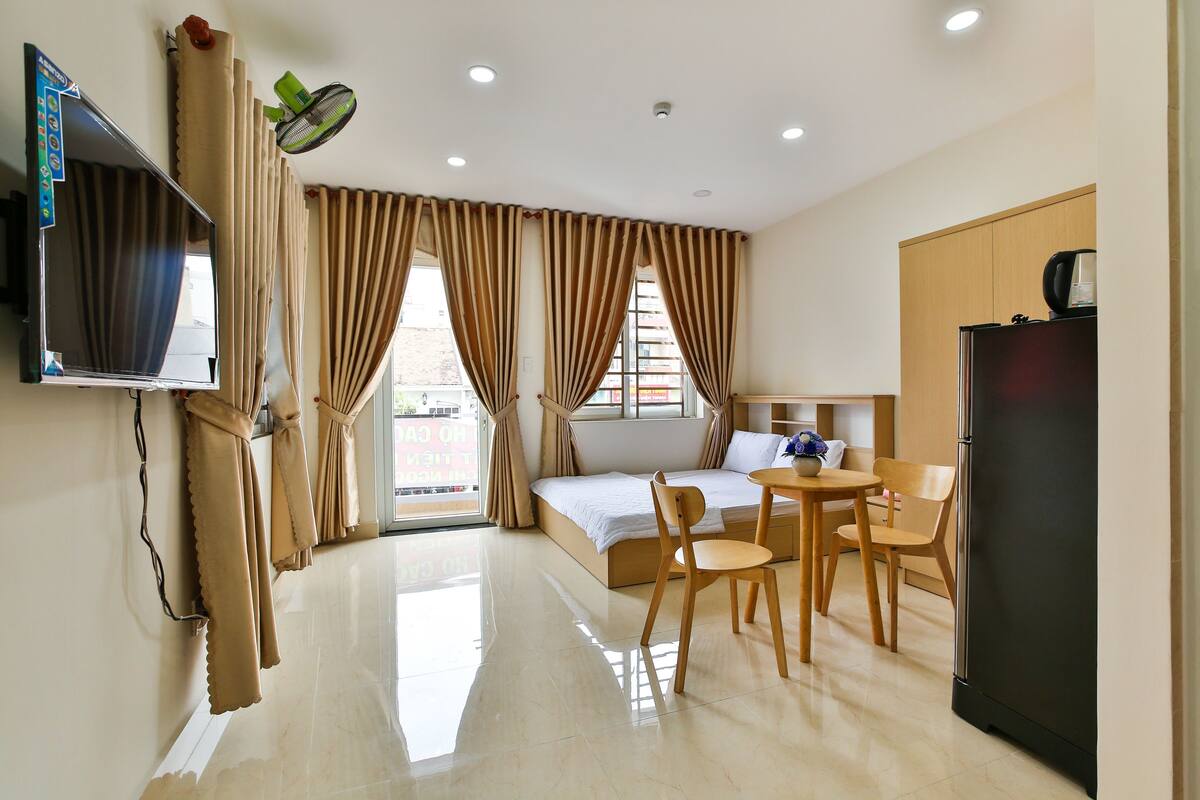 A hotel room with a bed, a TV, a dining area with chairs, a refrigerator, and curtains.
