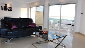 Una sala de estar moderna con un sofá de cuero negro, una mesa de centro de vidrio y un gran ventanal corredera que da a un balcón exterior con muebles blancos.