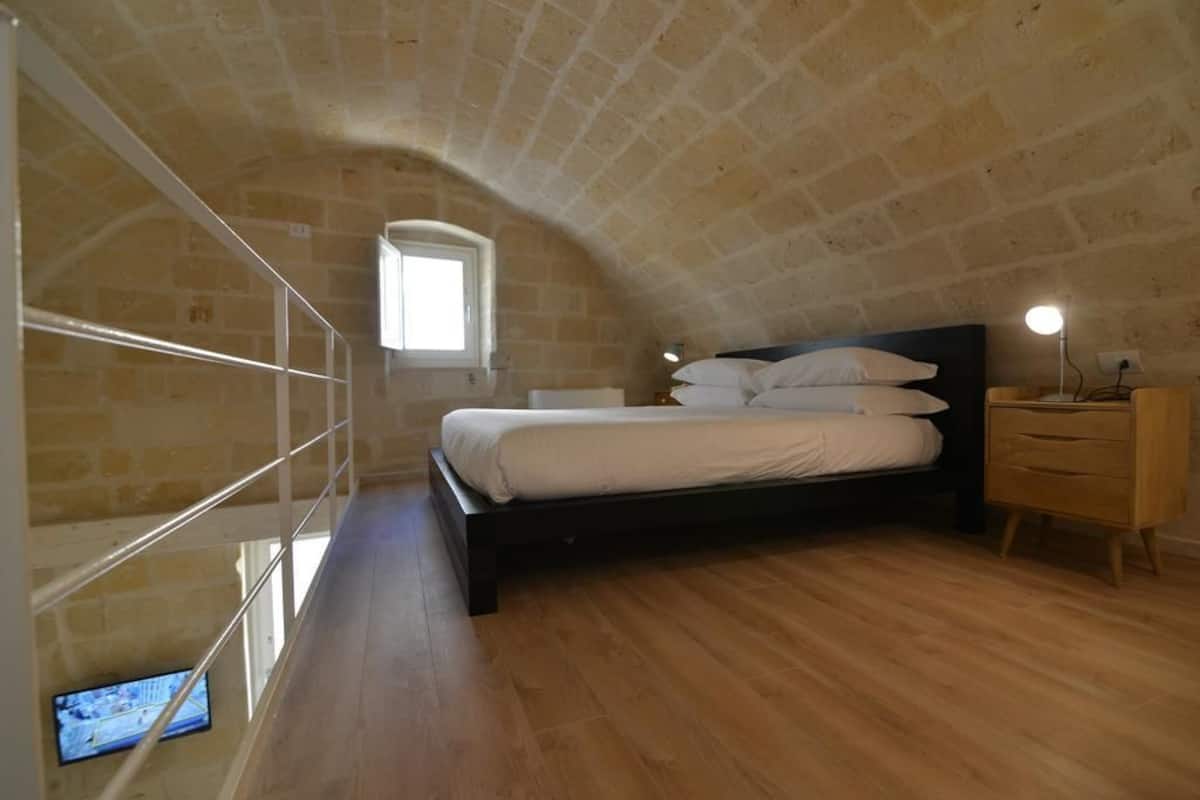 A bedroom with a sloped ceiling, a bed with white bedding, a wooden dresser, and a small bedside table with a lamp.
