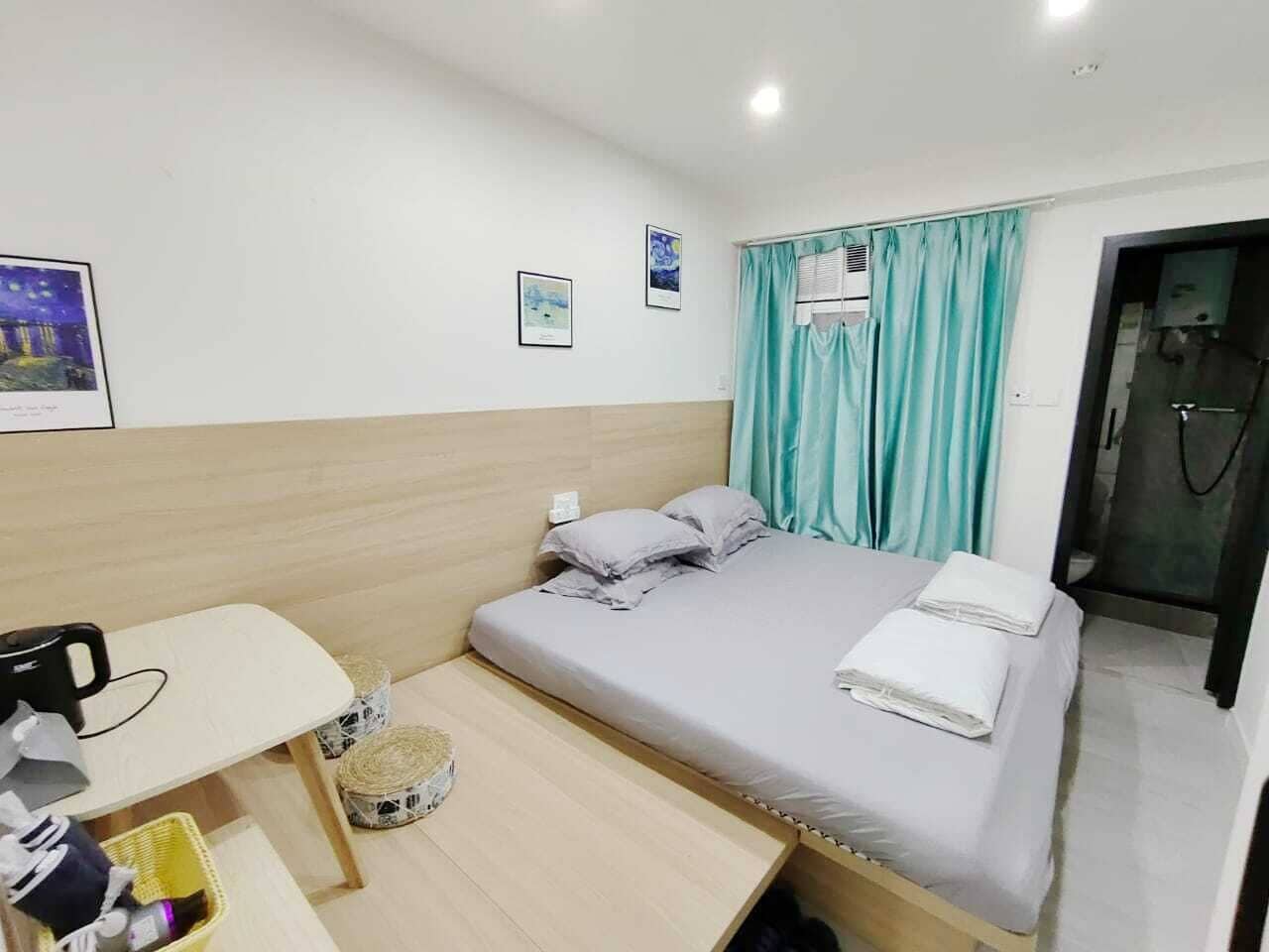 A small, neatly arranged hotel room with a bed, a desk, and a chair. There are framed pictures on the wall, a window with curtains, and a door leading to a bathroom.