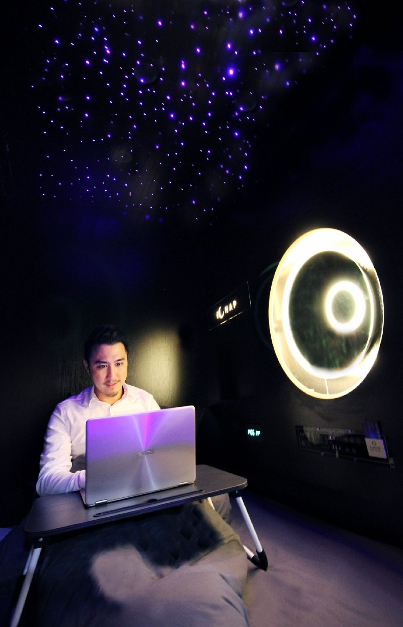 Uma pessoa sentada em uma cama com um laptop, uma luminária circular grande e um teto estrelado.