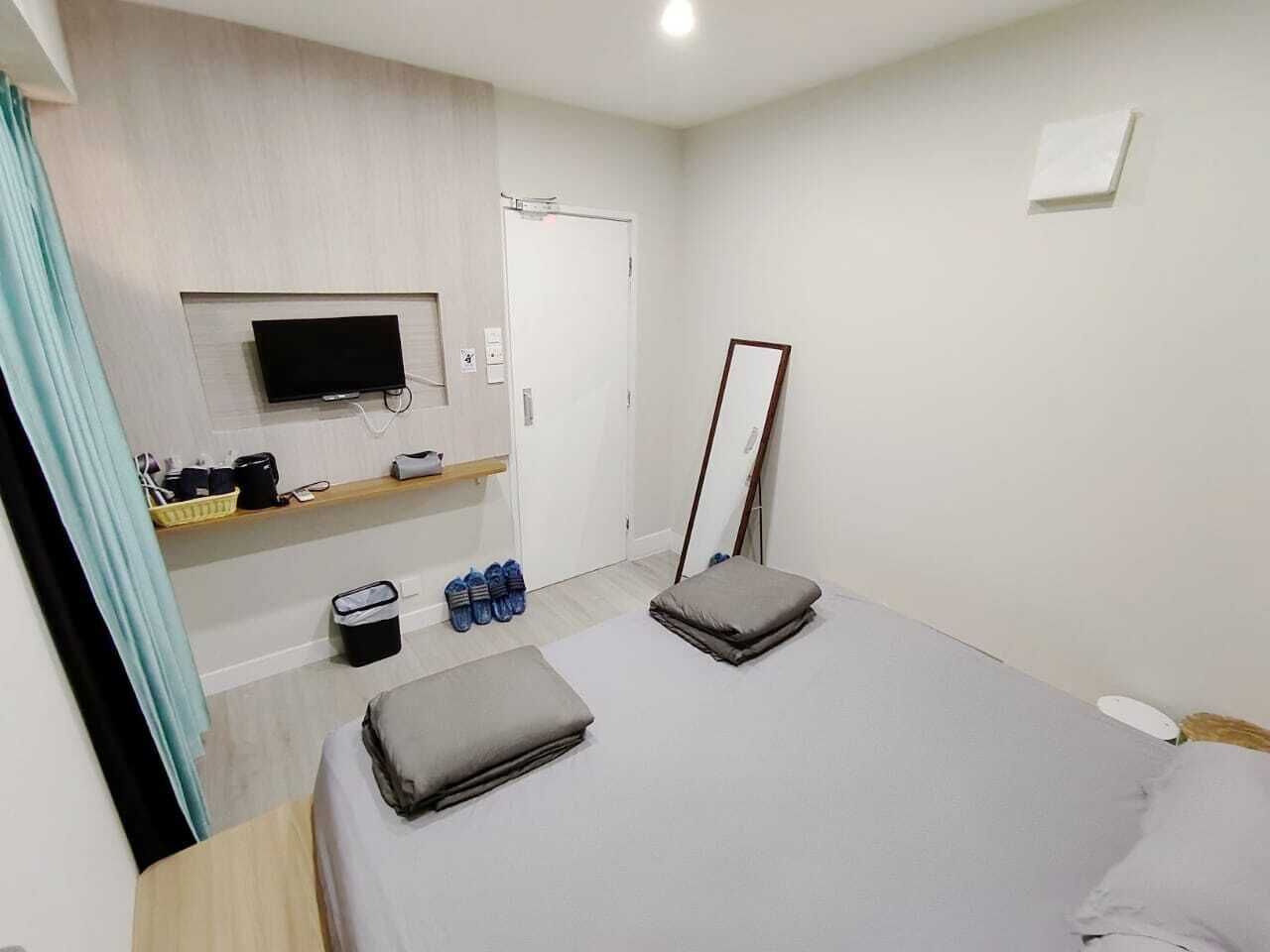 A modern hotel room with a bed, a TV mounted on the wall, a small shelf with toiletries, and a door.