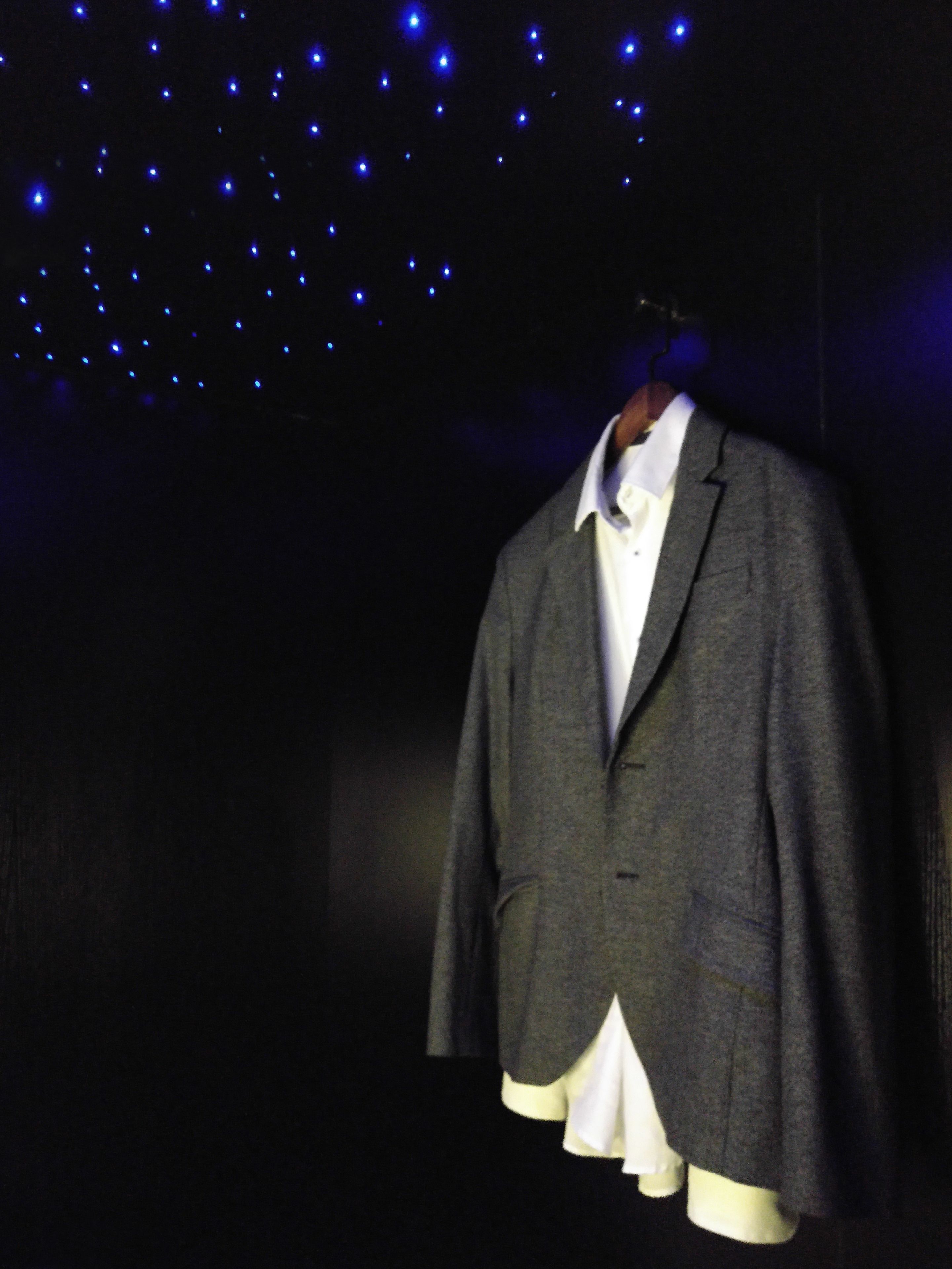 Un maniquí vestido con un blazer gris y una camisa blanca, colgado sobre un fondo oscuro con pequeñas luces azules.