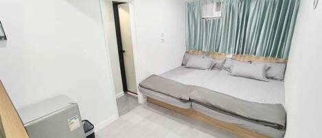 A small, modern bedroom with a bed, a small cabinet, and a door leading to another room.