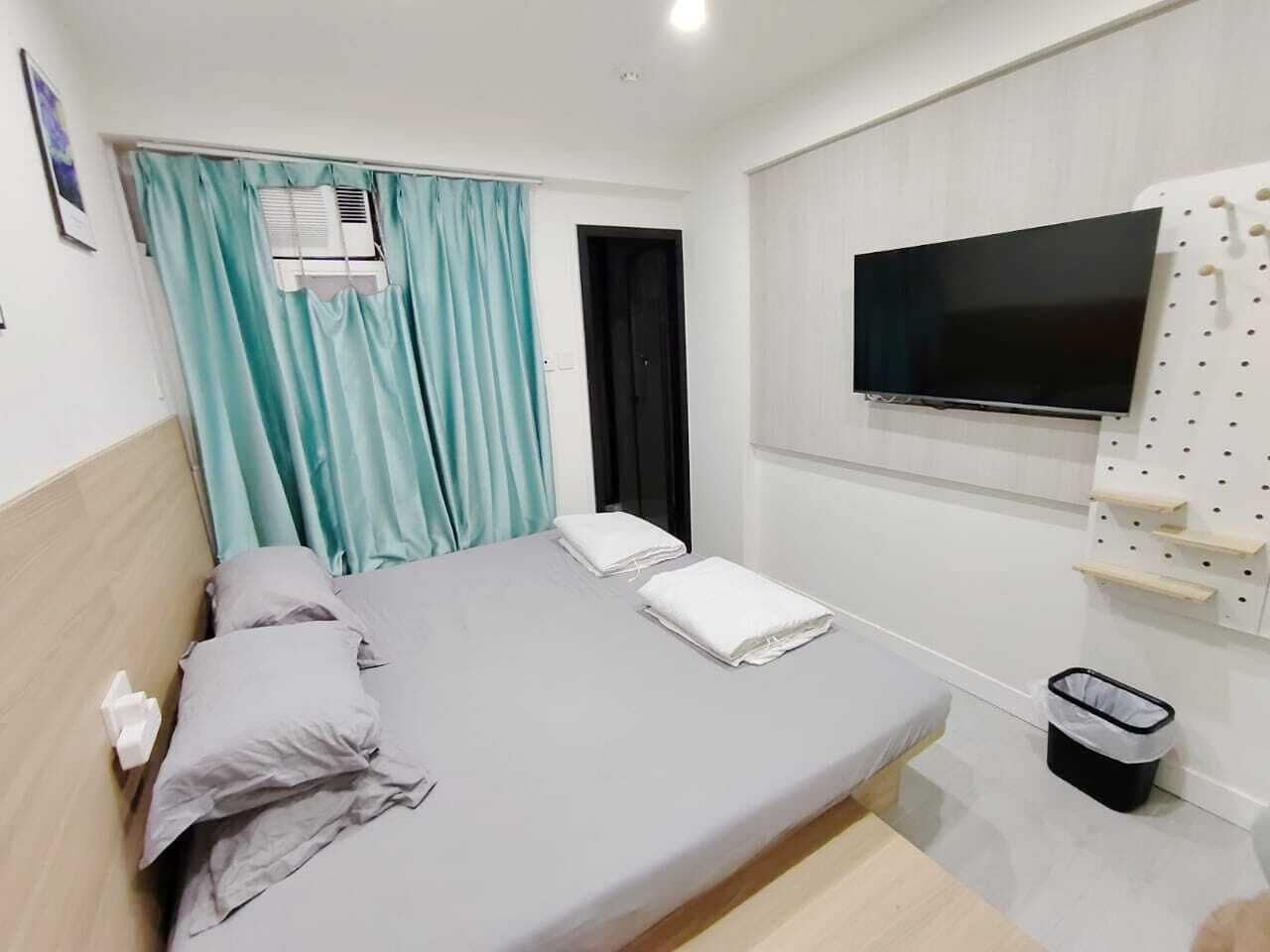 Habitación con cama individual, colchón gris, cortinas verde azulado, televisor montado y estante con ganchos.