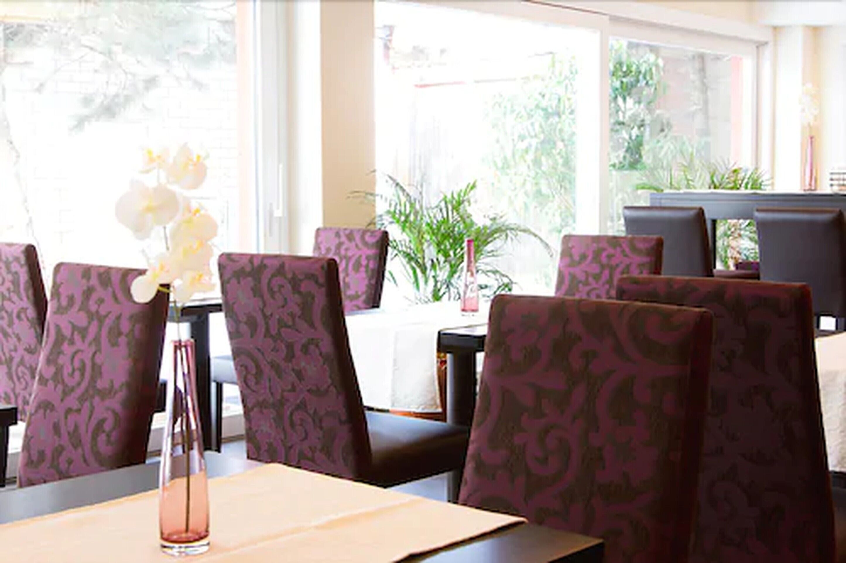 A restaurant interior with patterned chairs, a table with a vase of flowers, and large windows with a view of greenery outside.