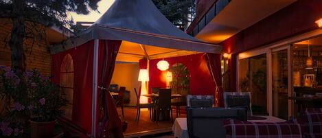 A tented outdoor dining area with tables and chairs, a red wall, and a glass door leading inside.