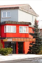A two-story building with a red and grey facade, a visible entrance, and a small tree to the right.
