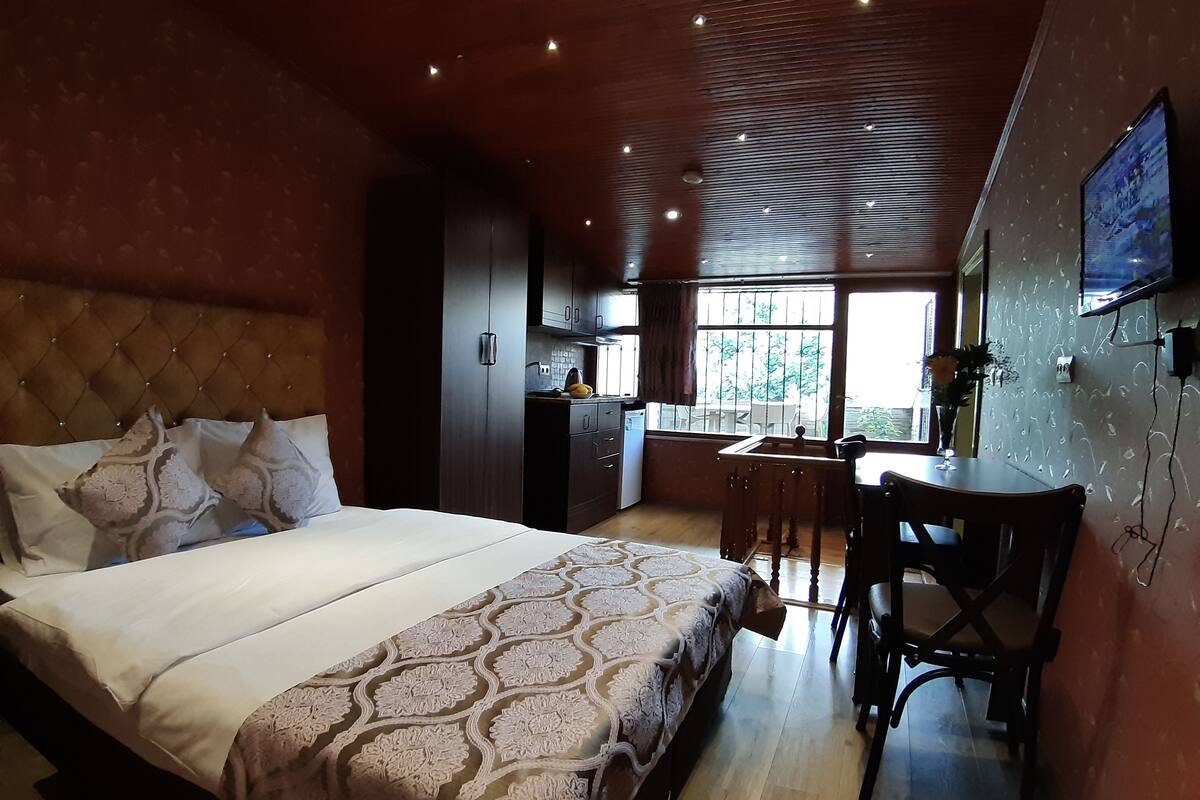 A bedroom with a large bed, a wooden ceiling, a small kitchen area, and a TV mounted on the wall.