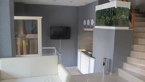 A modern living room with a white sofa, a flat-screen TV mounted on the wall, a staircase with a fish tank, and a small kitchen area. - Idrisoglu Hotel (Kastamonu)