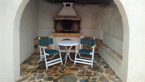 Eine Terrasse im Freien mit Steinboden, zwei weiße Stühle mit grünen Kissen, ein weißer Tisch und ein Steinkamin.