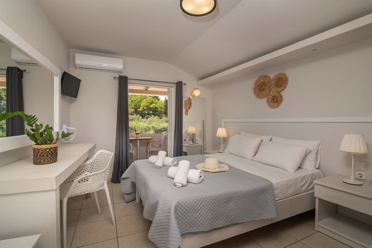 A modern hotel room with a bed, a desk, a chair, a TV, and a view of greenery outside.