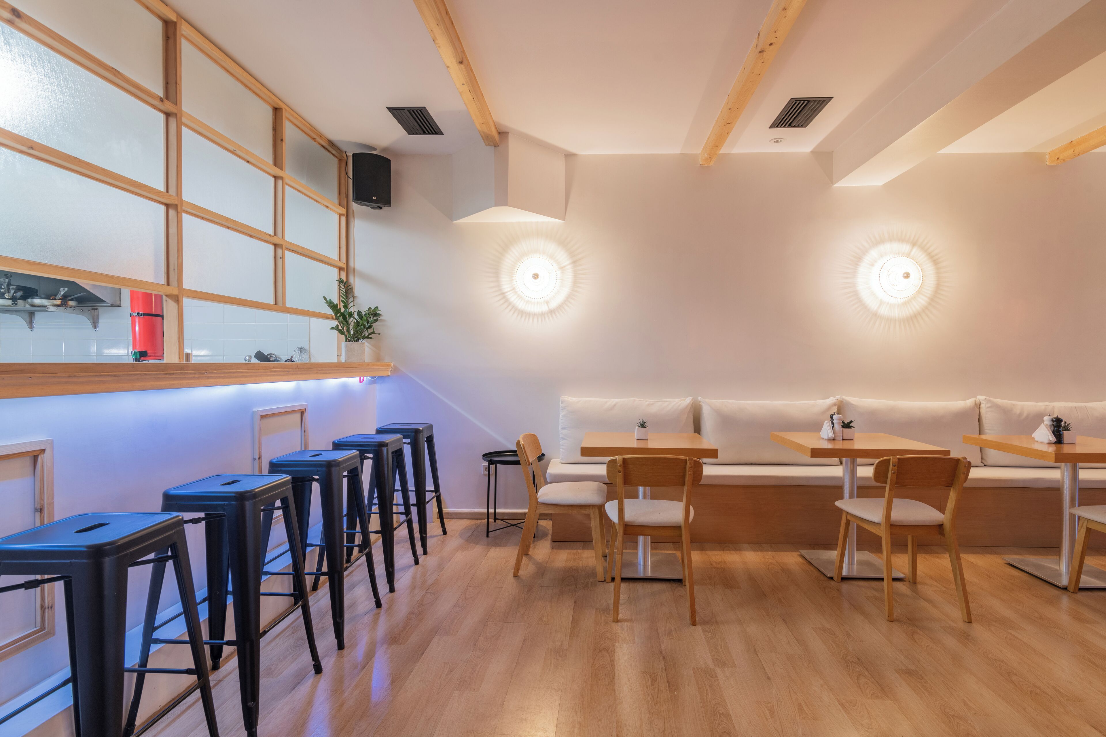 A modern restaurant interior with wooden beams, a long counter with stools, and a seating area with tables and chairs.