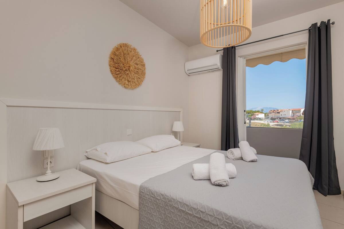 A bedroom with a bed, a bedside table with a lamp, a wall-mounted air conditioner, and a view of the outside through a window with curtains.