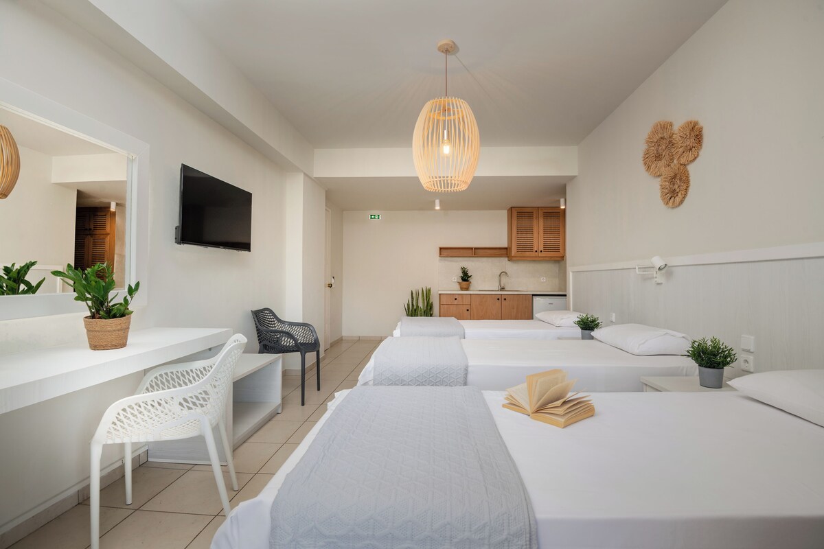 A modern hotel room with a white color scheme, a flat-screen TV, a dining area, and a kitchenette.