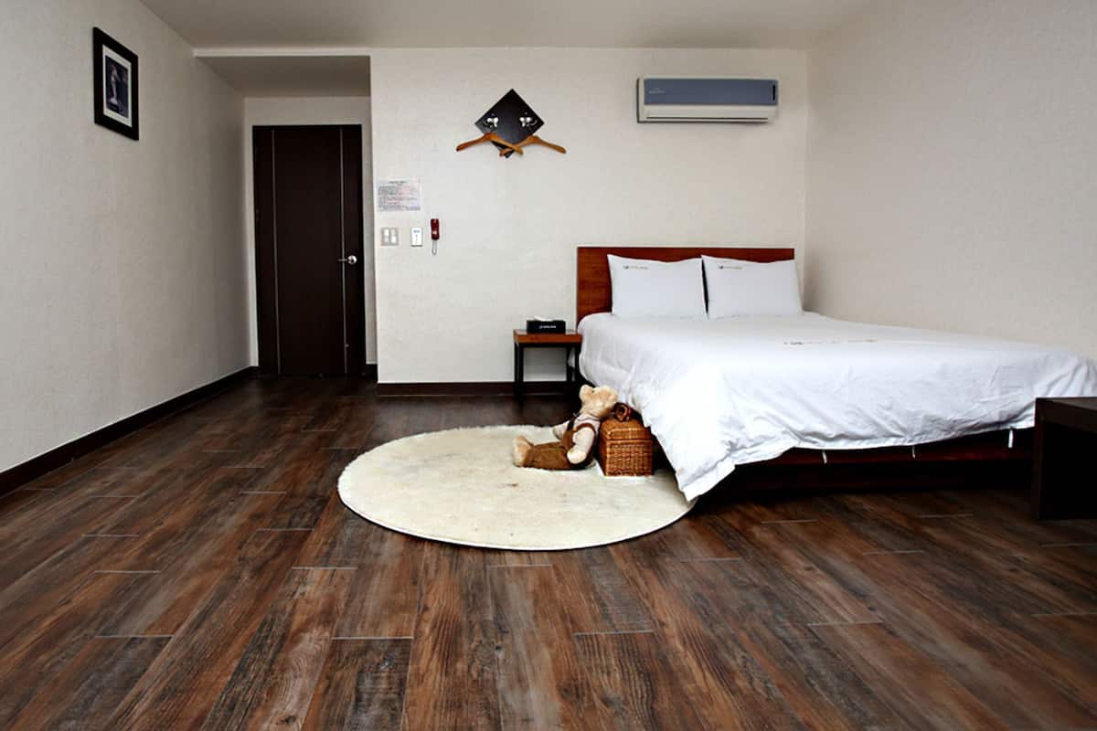 Habitación de hotel con piso de madera, cama con ropa blanca, mesita de noche, un oso de peluche y aire acondicionado instalado en la pared.