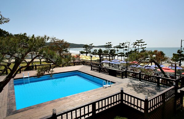 A swimming pool with a wooden deck and a wooden fence, surrounded by beach umbrellas and a view of the ocean. - Manripo Hotel (Taean)