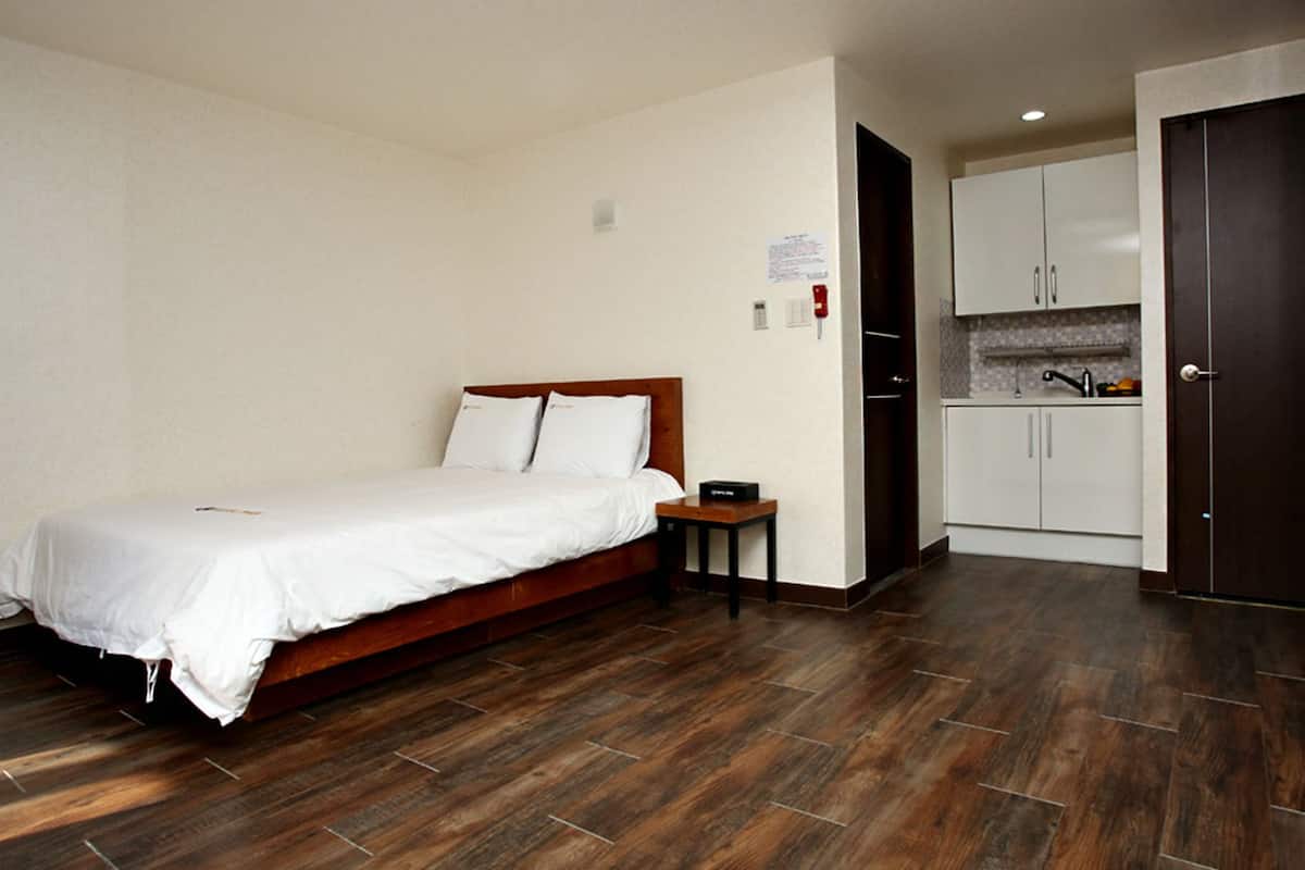 Habitación de hotel con cama de madera, una mesita de noche pequeña, una cocina integrada con armarios blancos y un piso de madera oscura.