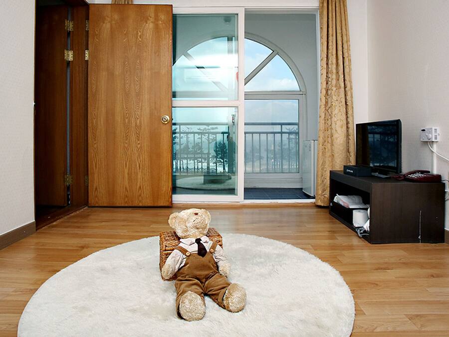 Quarto com piso de madeira, tapete branco e fofo, um urso de pelúcia, televisão sobre um suporte e uma porta de vidro deslizante que dá para uma varanda.