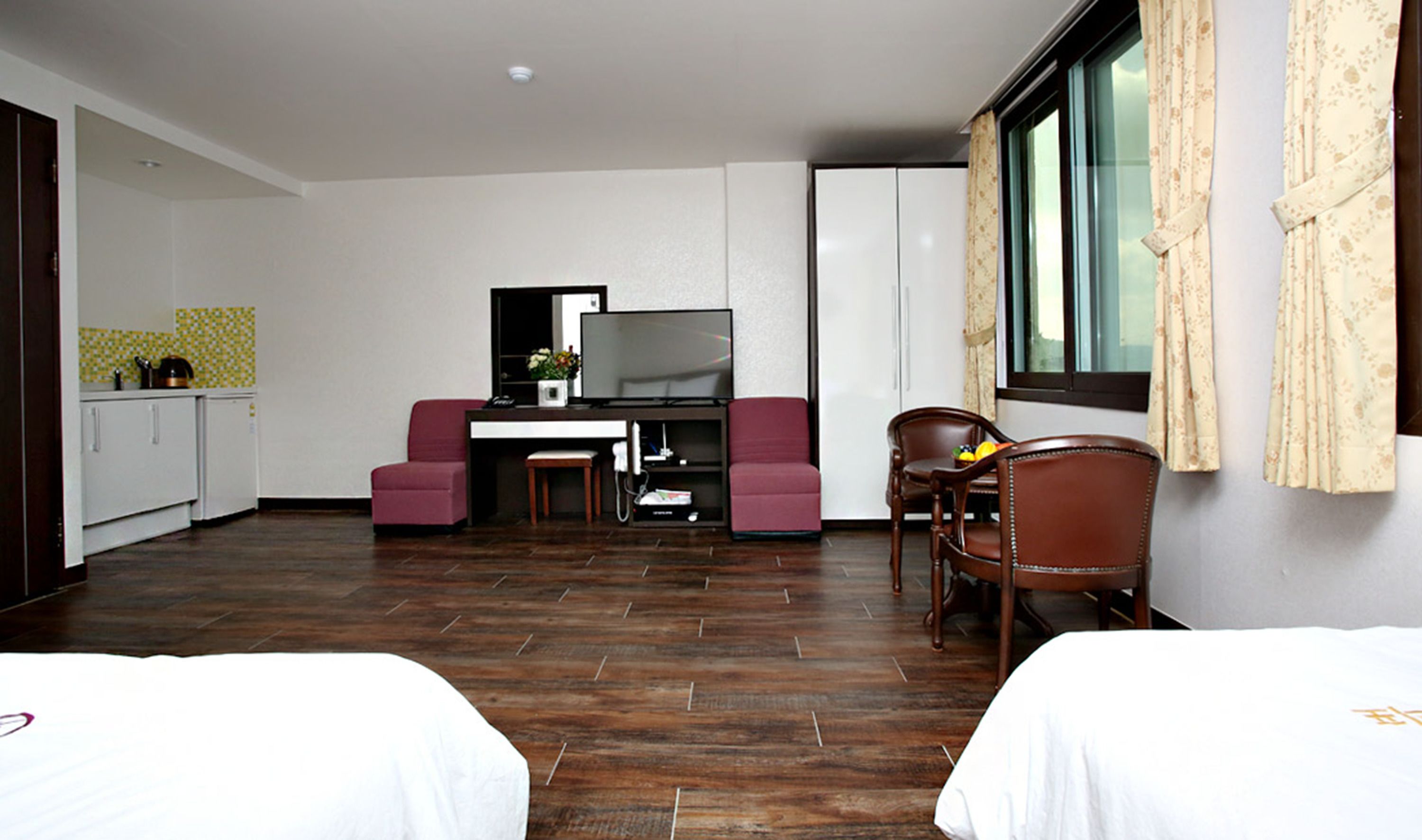 A hotel room with two beds, a desk with a TV, a small kitchenette, and a window with curtains.