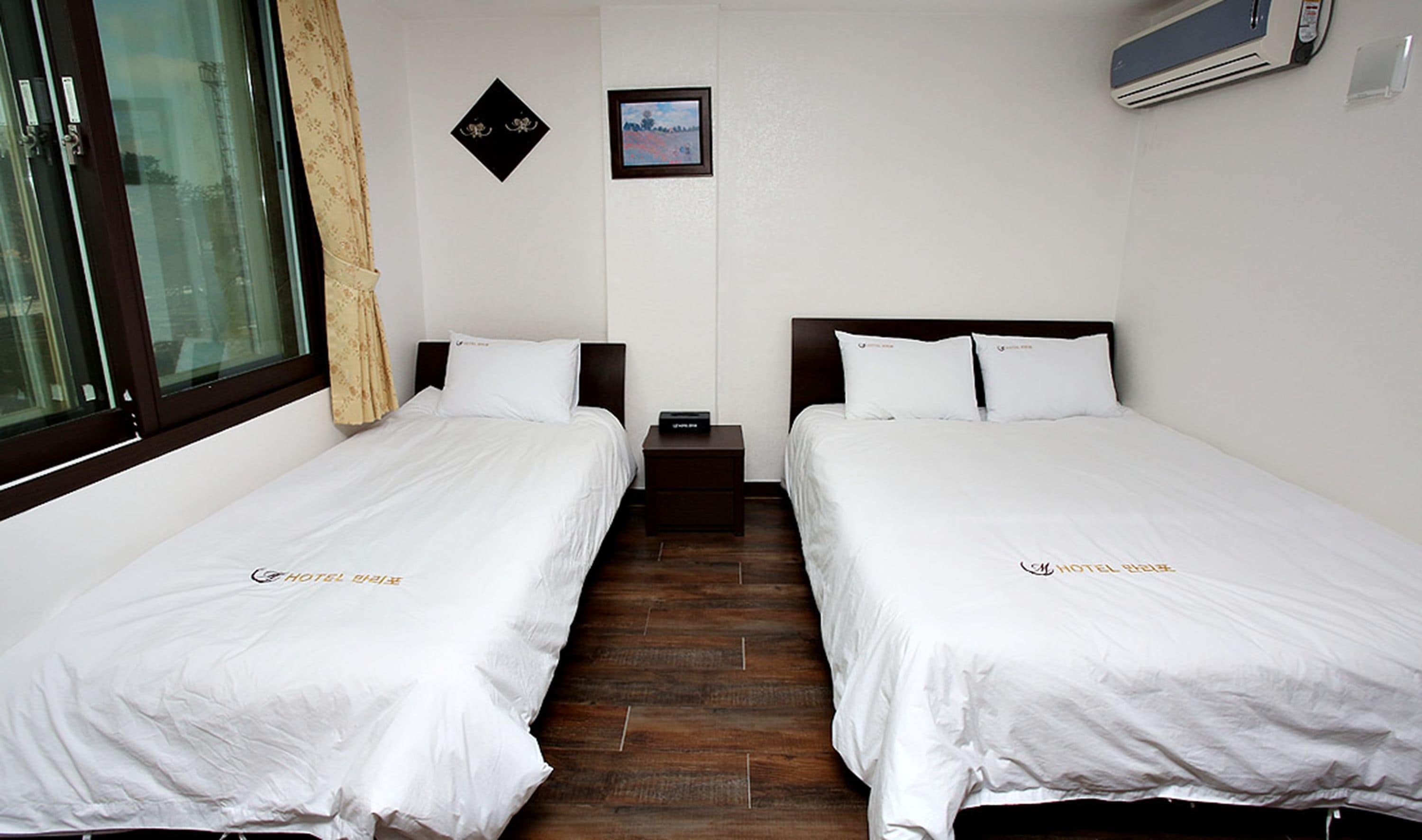 Habitación de hotel con dos camas individuales, una mesita de noche, una pequeña mesa, una ventana con cortina y un aire acondicionado instalado en la pared.