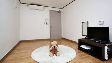 Habitació amb sòl de fusta, una catifa blanca, un os de peluix, un televisor sobre un suport i una porta amb interruptor de llum i detector de fum.