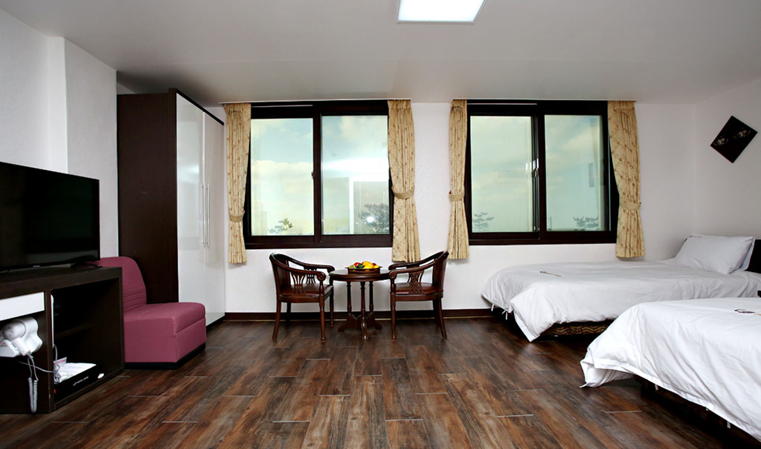 A hotel room with a TV, a pink chair, a dining table with chairs, and two beds.