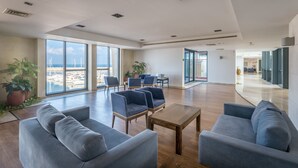 Un vestíbulo espacioso con diseño moderno, que cuenta con una amplia zona de estar con sofás y sillones, una mesa de centro de madera y grandes ventanales con vistas a la playa y el puerto deportivo.