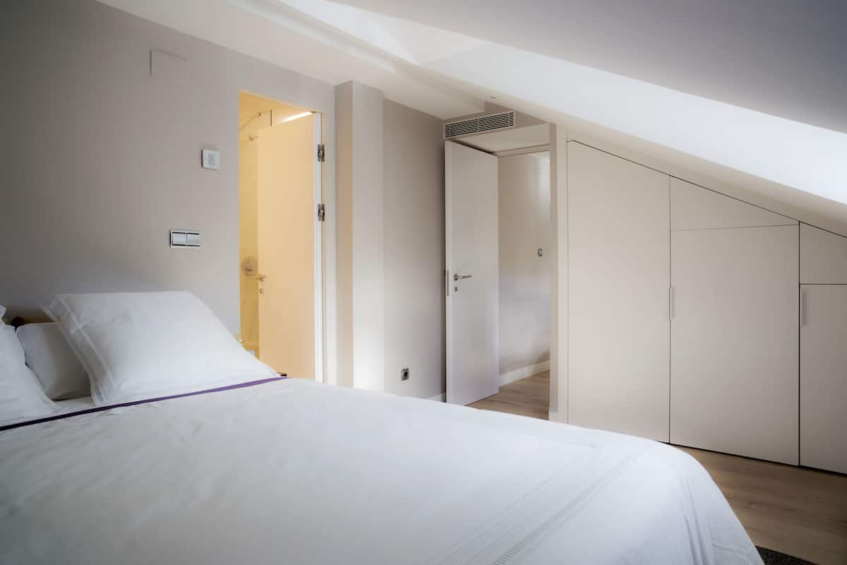 Un dormitorio con cama, paredes blancas, una puerta que da a un pasillo y un armario.