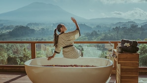 Mulher de vestido estampado sentada em uma banheira grande cheia de pétalas de rosa, apreciando a vista de uma paisagem montanhosa a partir de uma varanda com guarda-corpos de madeira.