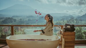 Mulher num vestido estampado sentada numa banheira grande cheia de água e pétalas de rosa, com vista para montanhas ao fundo.