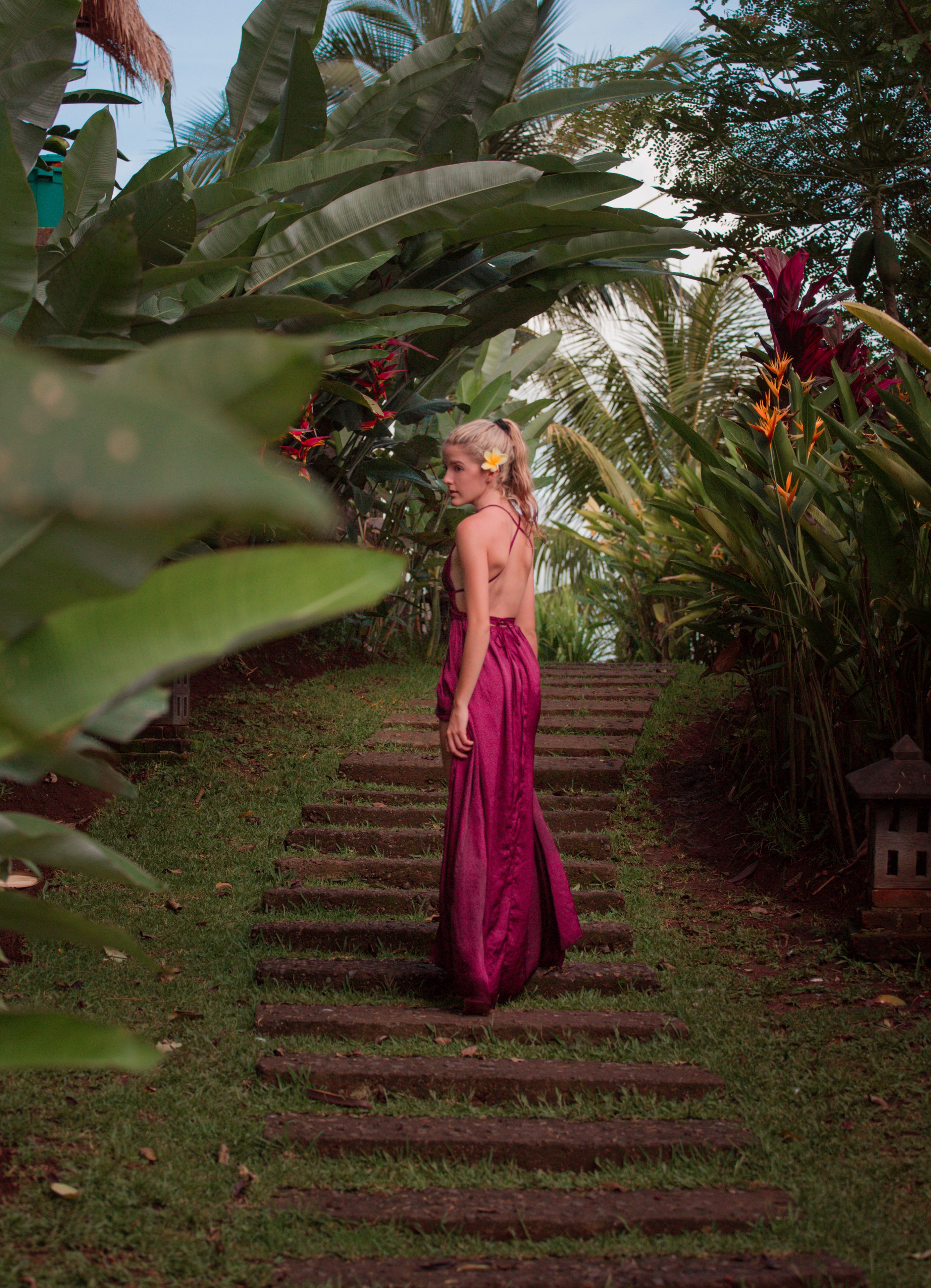 Una mujer con vestido rosa caminando por un sendero de piedra rodeado de exuberante follaje y plantas tropicales.