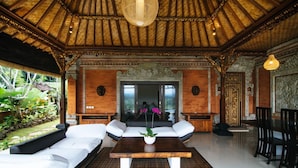 Um quarto espaçoso com teto de madeira, área de estar central com sofá e mesa de centro, e vista para um jardim exuberante através de uma porta de vidro.